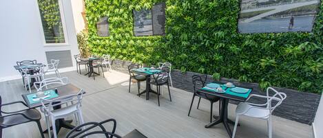 An outdoor dining area with tables and chairs, a green wall with plants, and a view of a river.