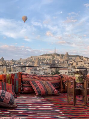 A rooftop terrace with colorful cushions, a view of a cityscape with unique rock formations, and a hot air balloon in the sky. - Kayra Cave Suites (Nevsehir)
