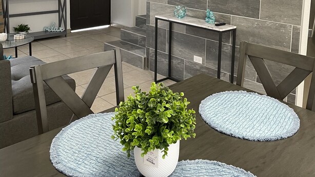 Un salon moderne avec une table à manger en bois foncé, des murs recouverts de carreaux gris et blancs, un téléviseur fixé au mur et un escalier menant à un autre niveau.