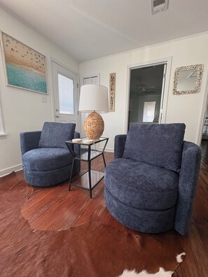 Un soggiorno con due poltrone blu, un tavolino in vetro, una lampada intrecciata, un tappeto in pelle di vacca e una parete con un cartello che riporta la scritta 'beach house'.