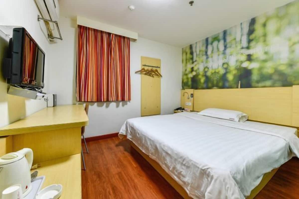 A hotel room with a bed, a desk with a kettle, a television, and a painting on the wall.