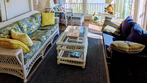 A room with a white wicker sofa, a blue sofa, a small table with magazines, and a view of a body of water through large windows. - Unique Waterfront Condo at the Boatyard! (Kennebunk)