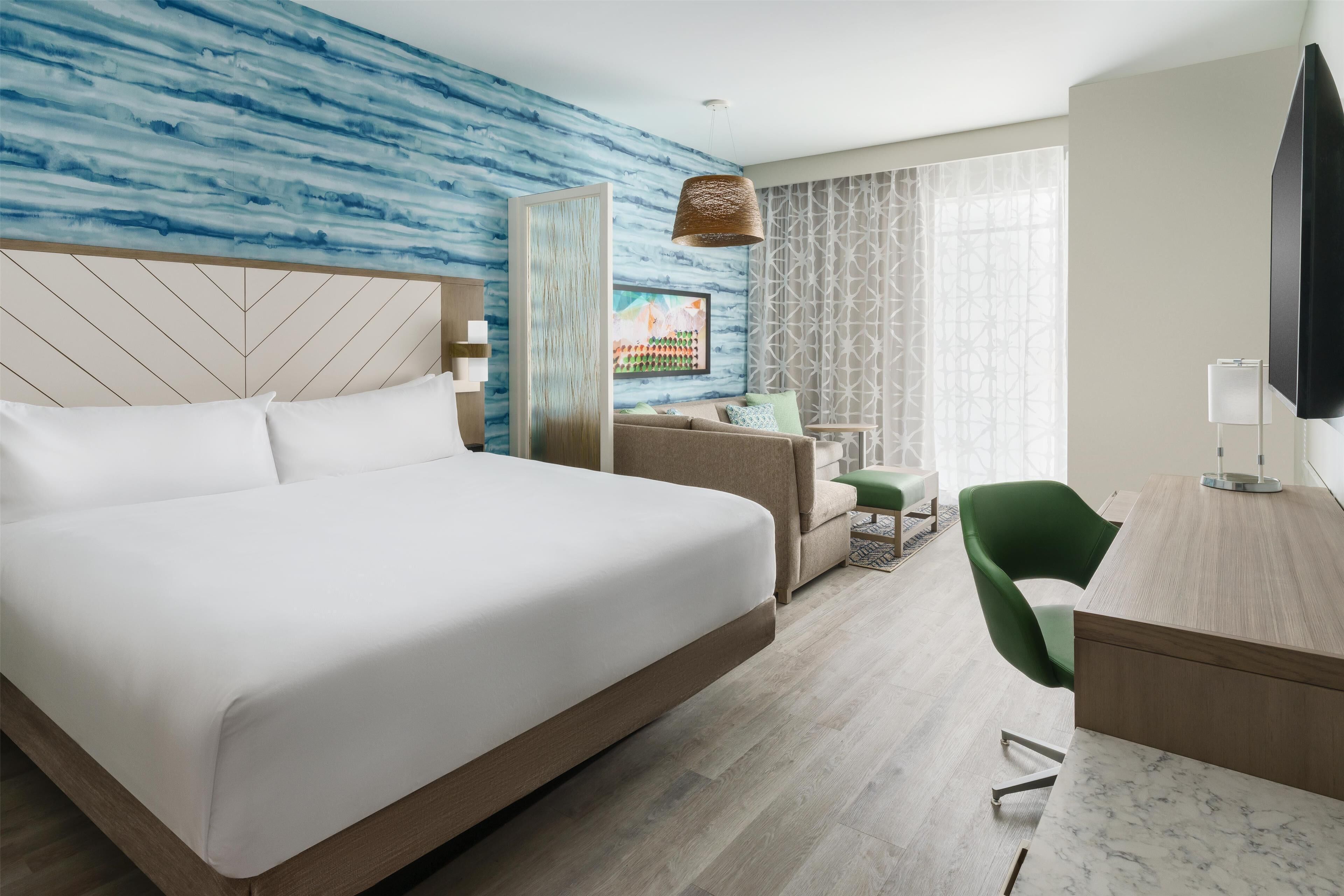 A modern hotel room with a large bed, a desk, a green chair, and a TV.