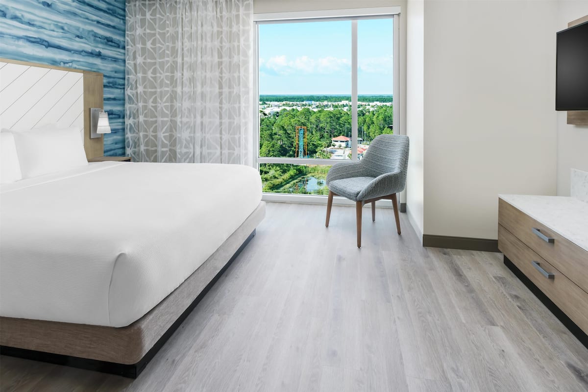 A modern hotel room with a large bed, a chair, a TV, and a window with a city view.