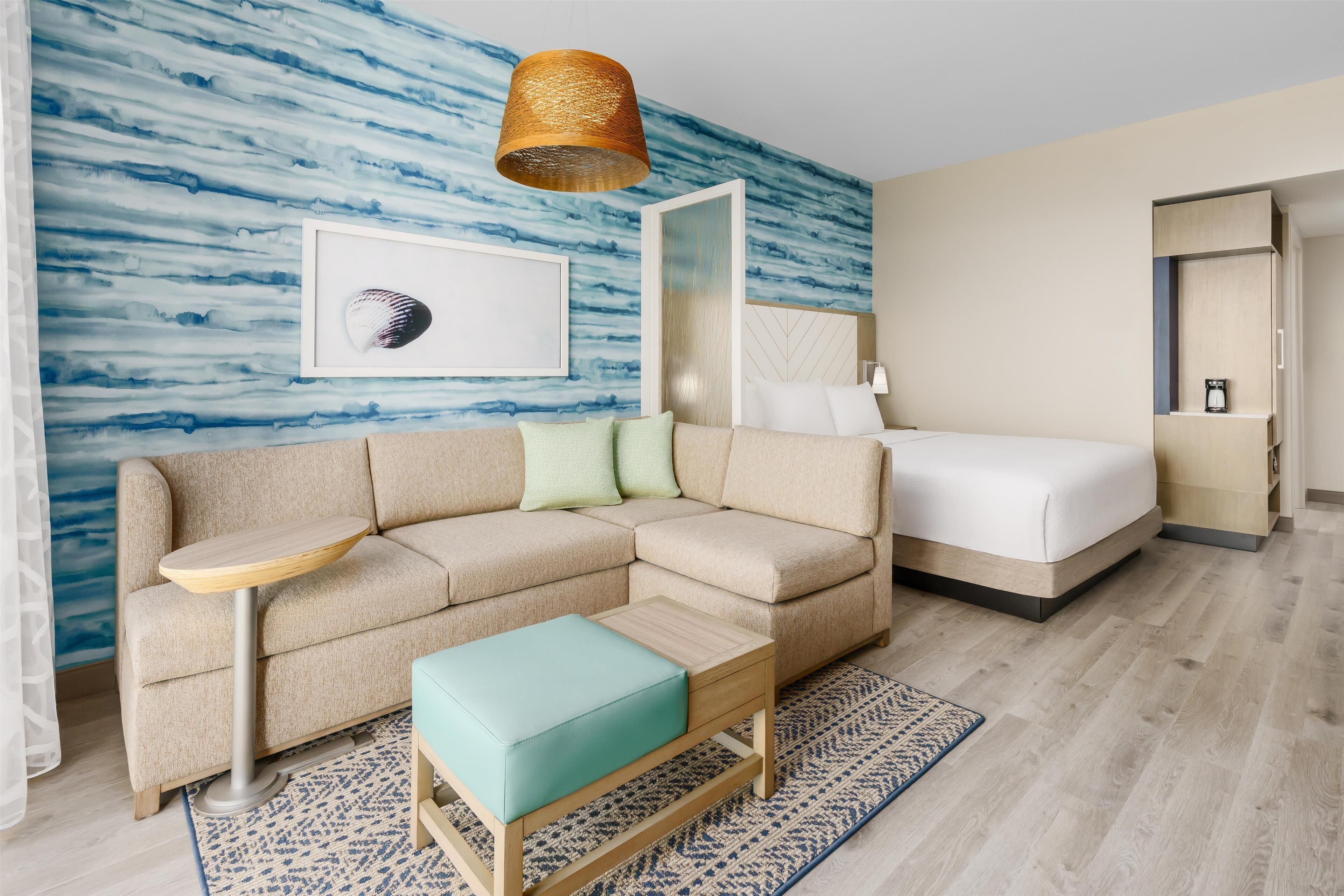 A modern hotel room with a sofa, a small table, a bed, a mirror, and a wall with a blue abstract design.