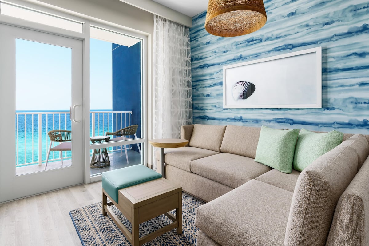 A living room with a beige sofa, a small wooden table, and a view of the ocean through a glass door.
