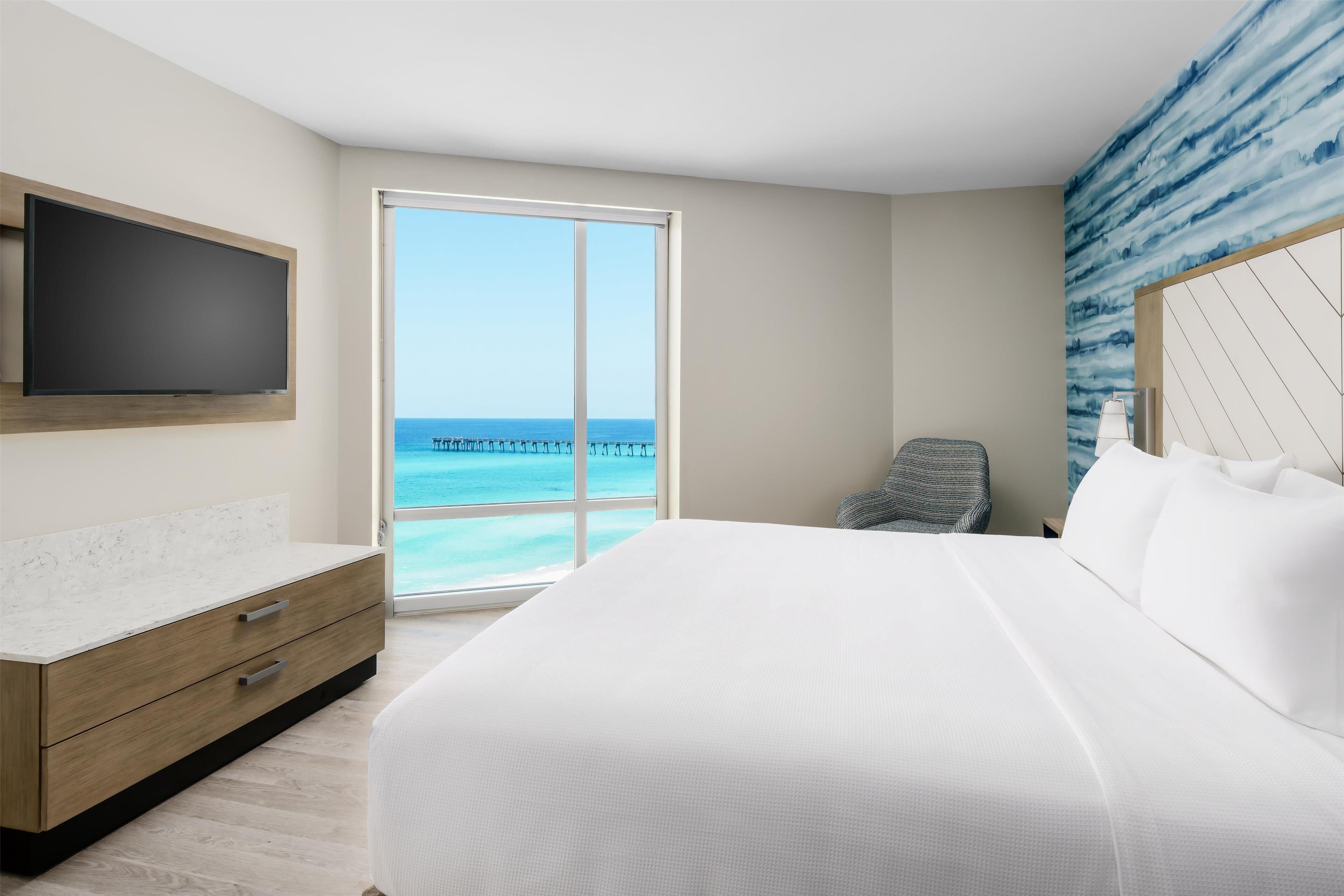 Ein Hotelzimmer mit einem großen Bett, einem an der Wand befestigten Fernseher, einer hölzernen Kommode und Blick aufs Meer.