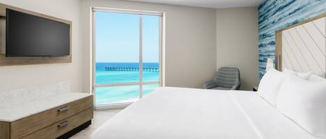 Habitación de hotel con una cama grande, un televisor montado en la pared, un aparador de madera y vista al océano.