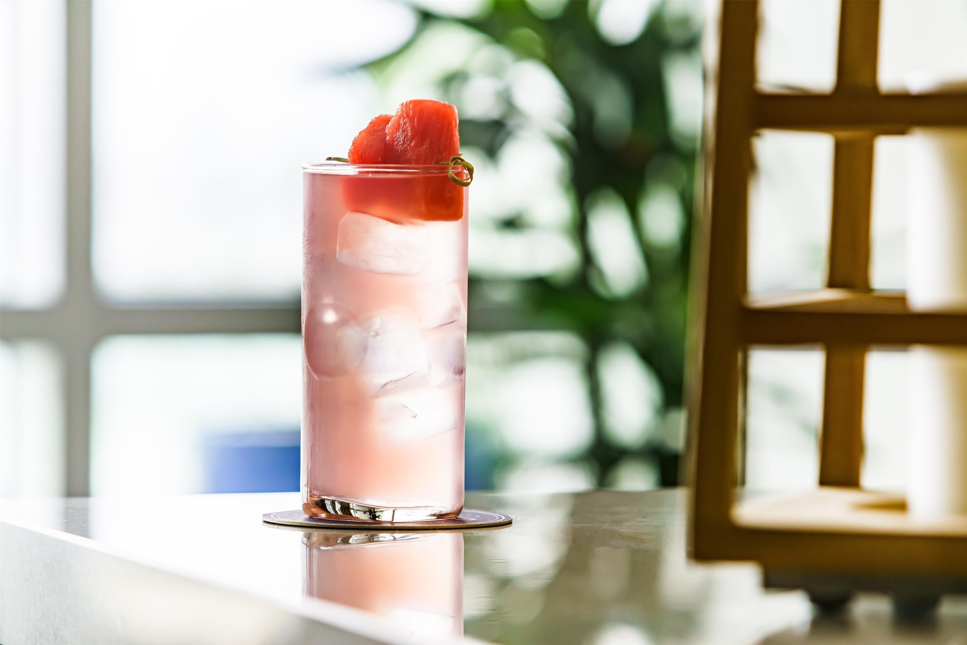 A tall glass with a pink beverage, ice cubes, and a slice of fruit on a reflective surface.