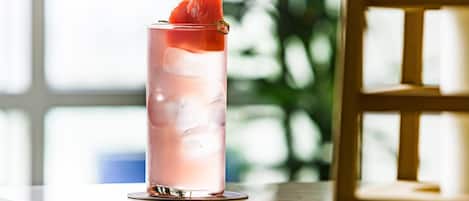 A tall glass with a pink beverage, ice cubes, and a slice of fruit on a reflective surface.
