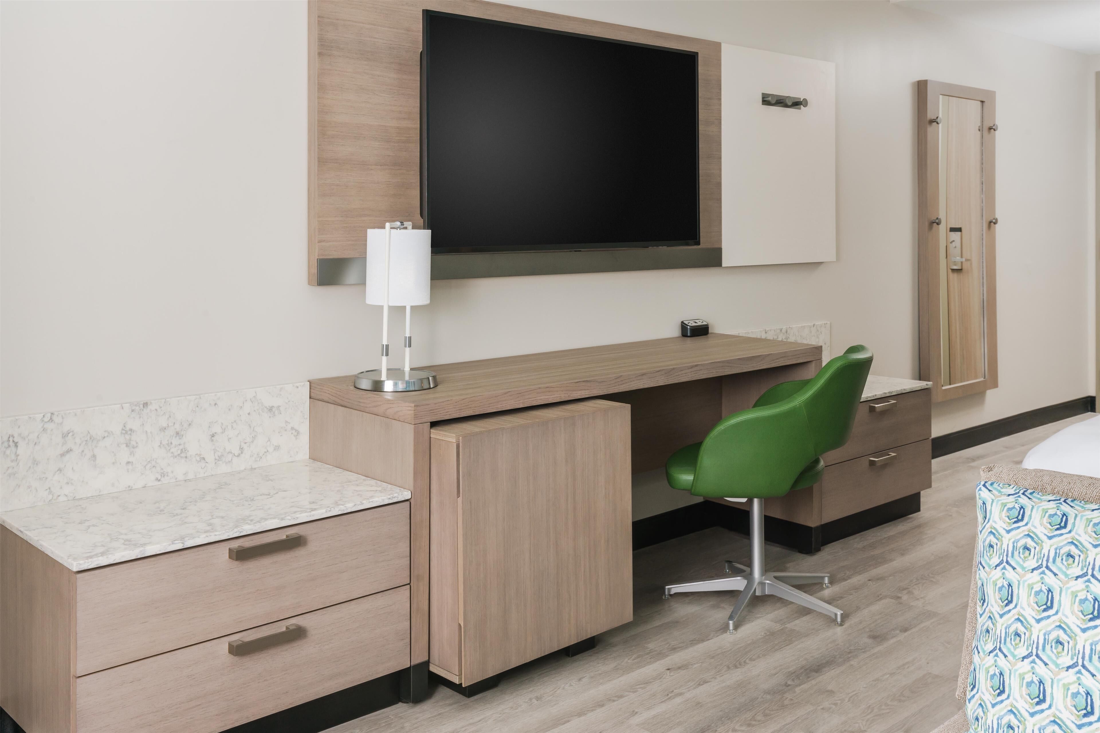 A modern hotel room with a wooden desk, a green chair, a flat-screen TV mounted on the wall, a bedside table, and a patterned throw on the bed.
