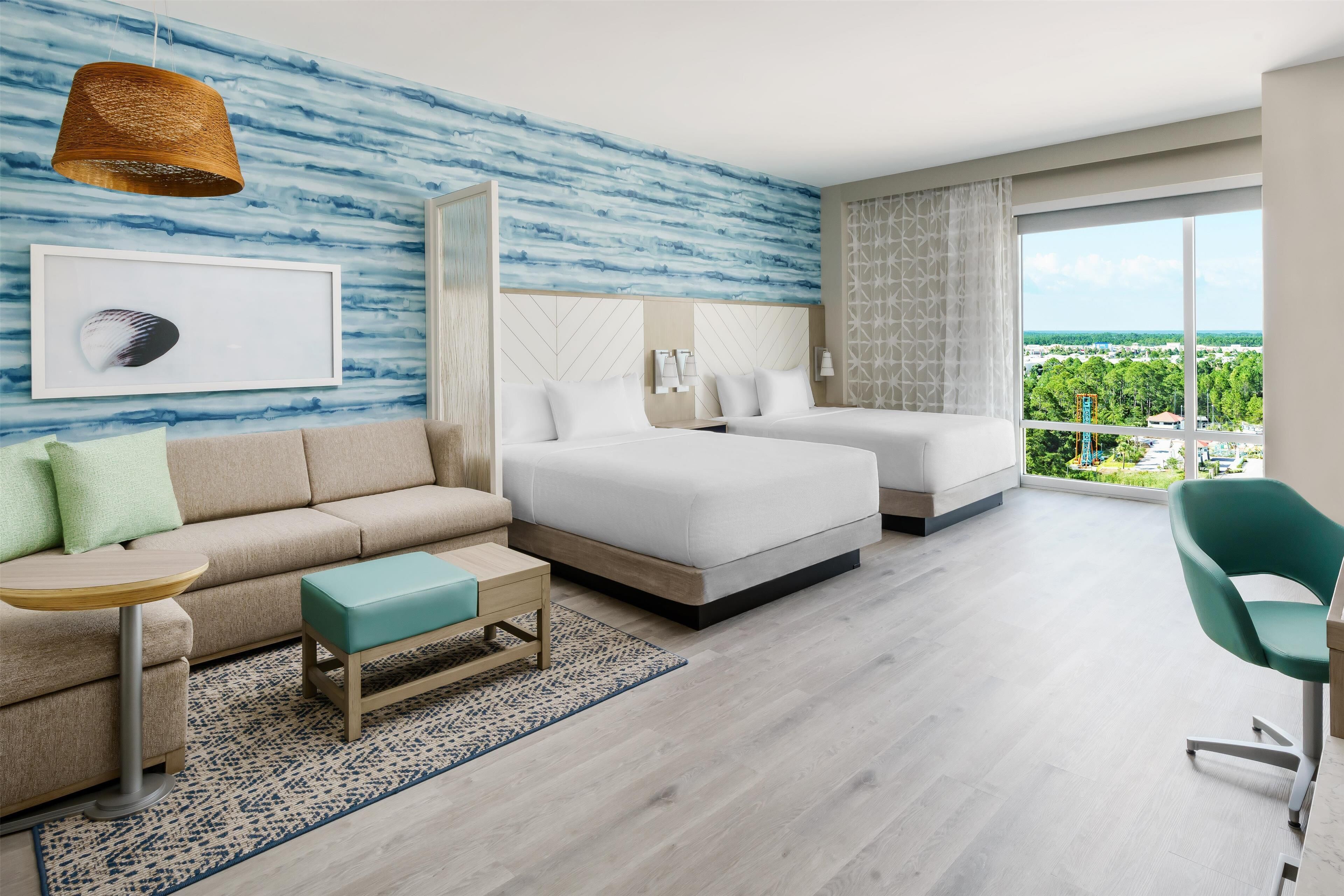 A modern hotel room with a large bed, a sofa, a small table, and a view of greenery outside.