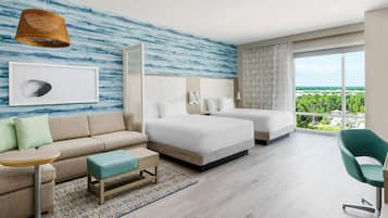A modern hotel room with a large bed, a sofa, a small table, and a view of greenery outside.