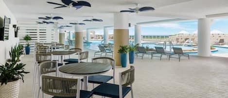 An indoor pool area with lounge chairs and tables, ceiling fans, and potted plants.
