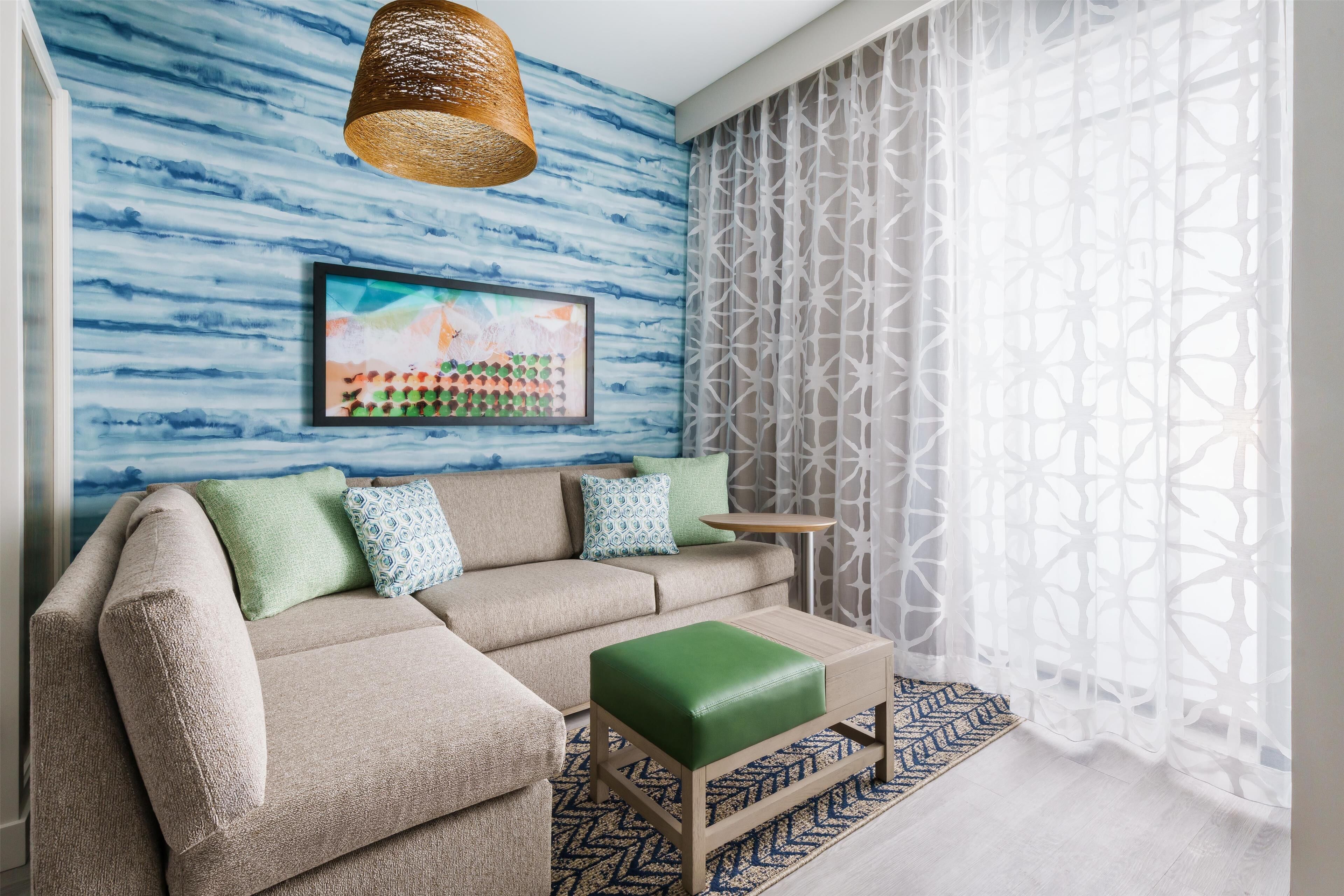 A living room with a sectional sofa, a green ottoman, a framed artwork, a woven light fixture, and a patterned window treatment.