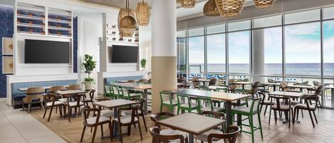 A spacious dining area with multiple tables and chairs, large windows offering a view of the sea, and wicker pendant lights.