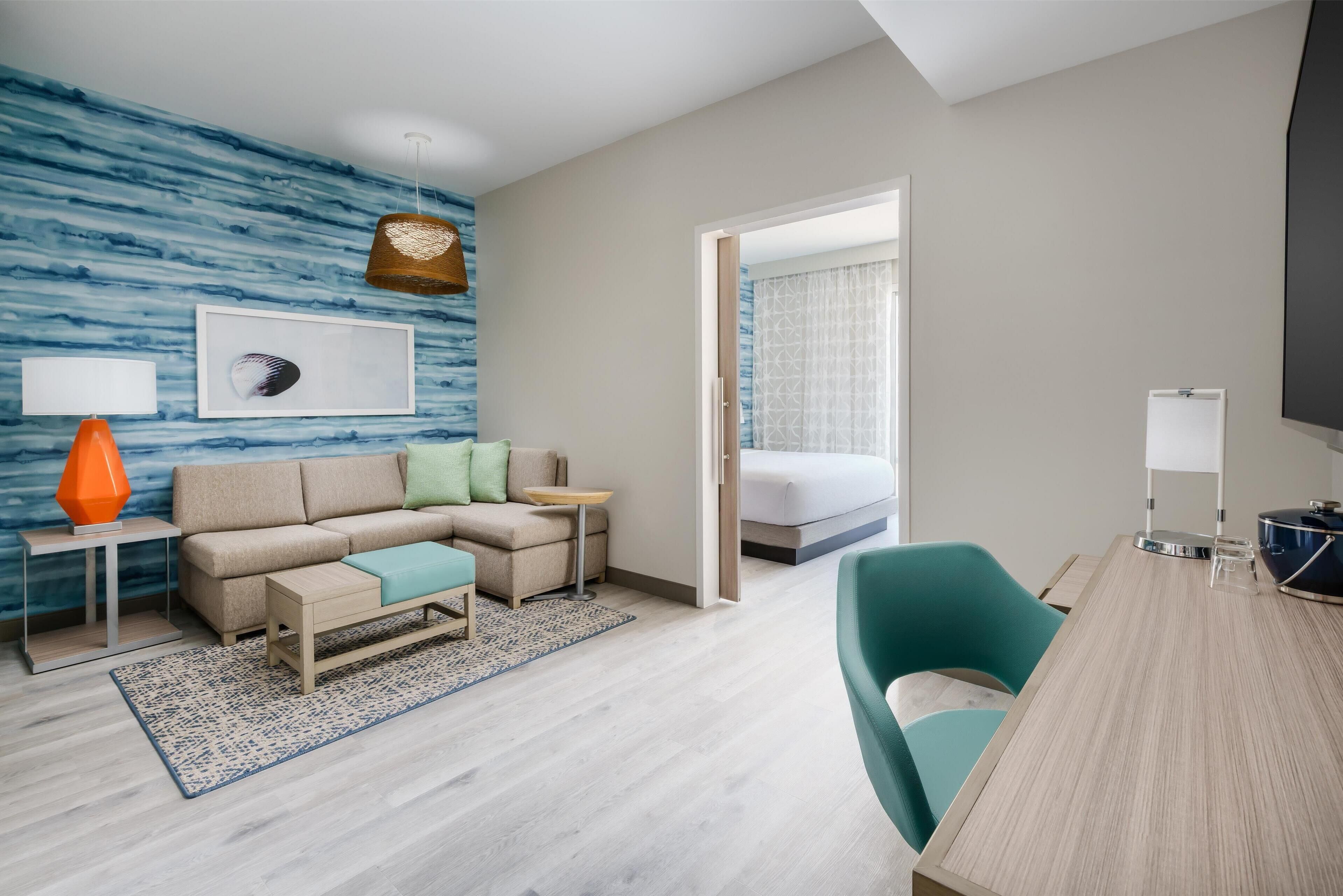 Una habitación de hotel moderna con un sofá modular, una mesita, un escritorio con una silla, una cama grande y vistas de la habitación contigua.
