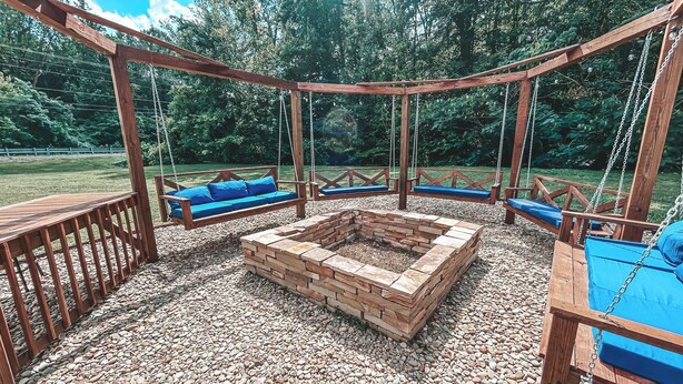Un ensemble de balançoires en bois avec des coussins bleus, un foyer central et un sol en gravier.