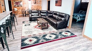 Un salon moderne avec un canapé d’angle en cuir, une table basse en bois et un tapis à motifs. On y trouve également un coin bar avec des tabourets et un luminaire suspendu.