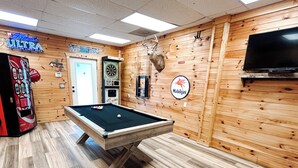 Une table de billard dans une pièce aux murs en bois, un jeu de fléchettes, une tête de cerf empaillée et un téléviseur.