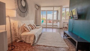Une pièce avec un canapé blanc, un lampadaire, un téléviseur fixé au mur et une vue sur l’extérieur grâce à une grande fenêtre.