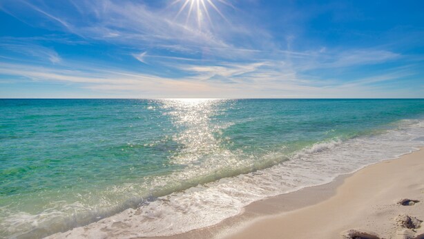 Uma praia com águas turquesa cristalinas, ondas suaves e um sol brilhante no céu.