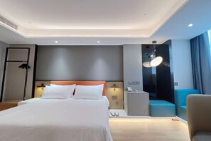 A modern hotel room with a large bed, a bedside table, a desk, and a seating area with a chair and a sofa.