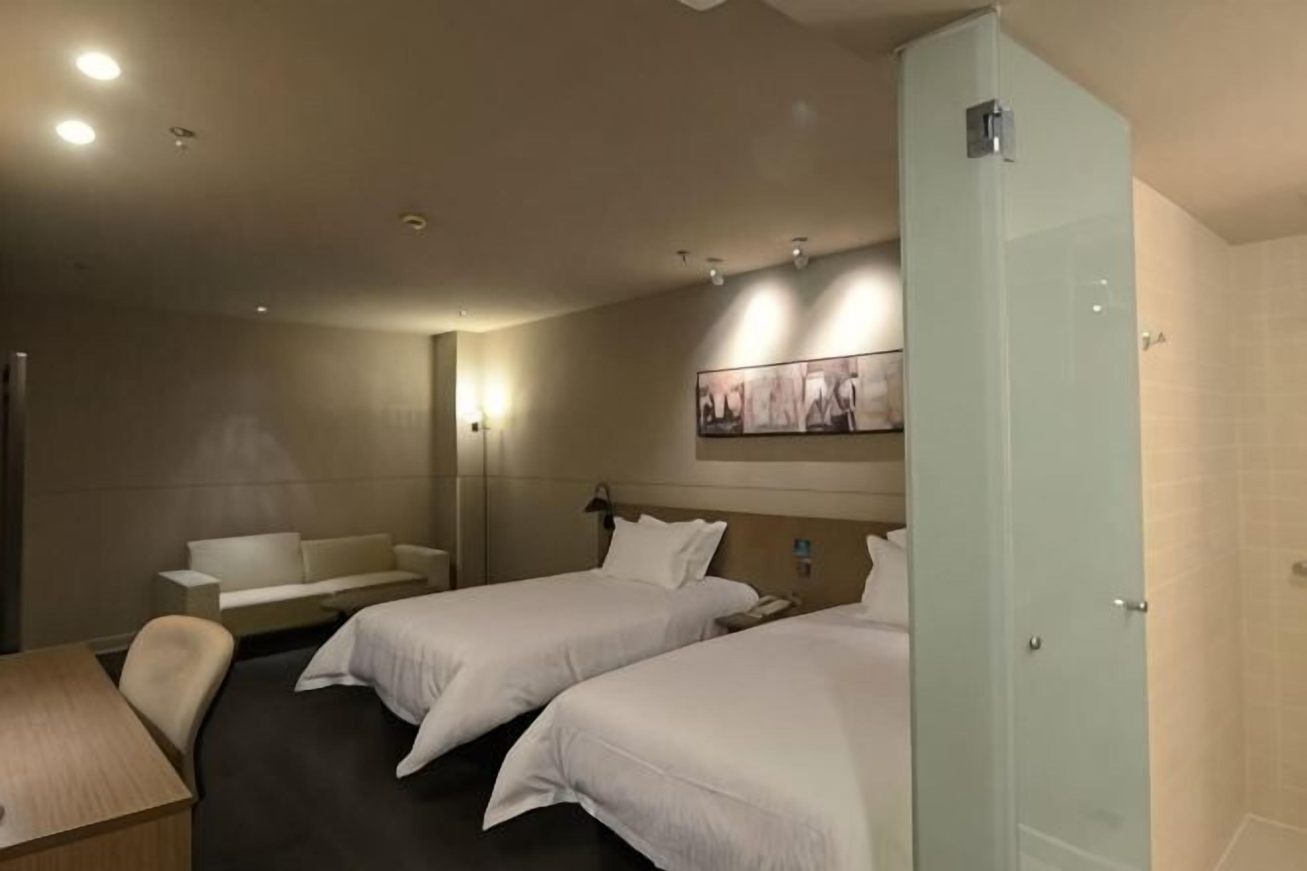 Hotellihuone, jossa on kaksi sänkyä, työpöytä, sohva ja suihkutila.