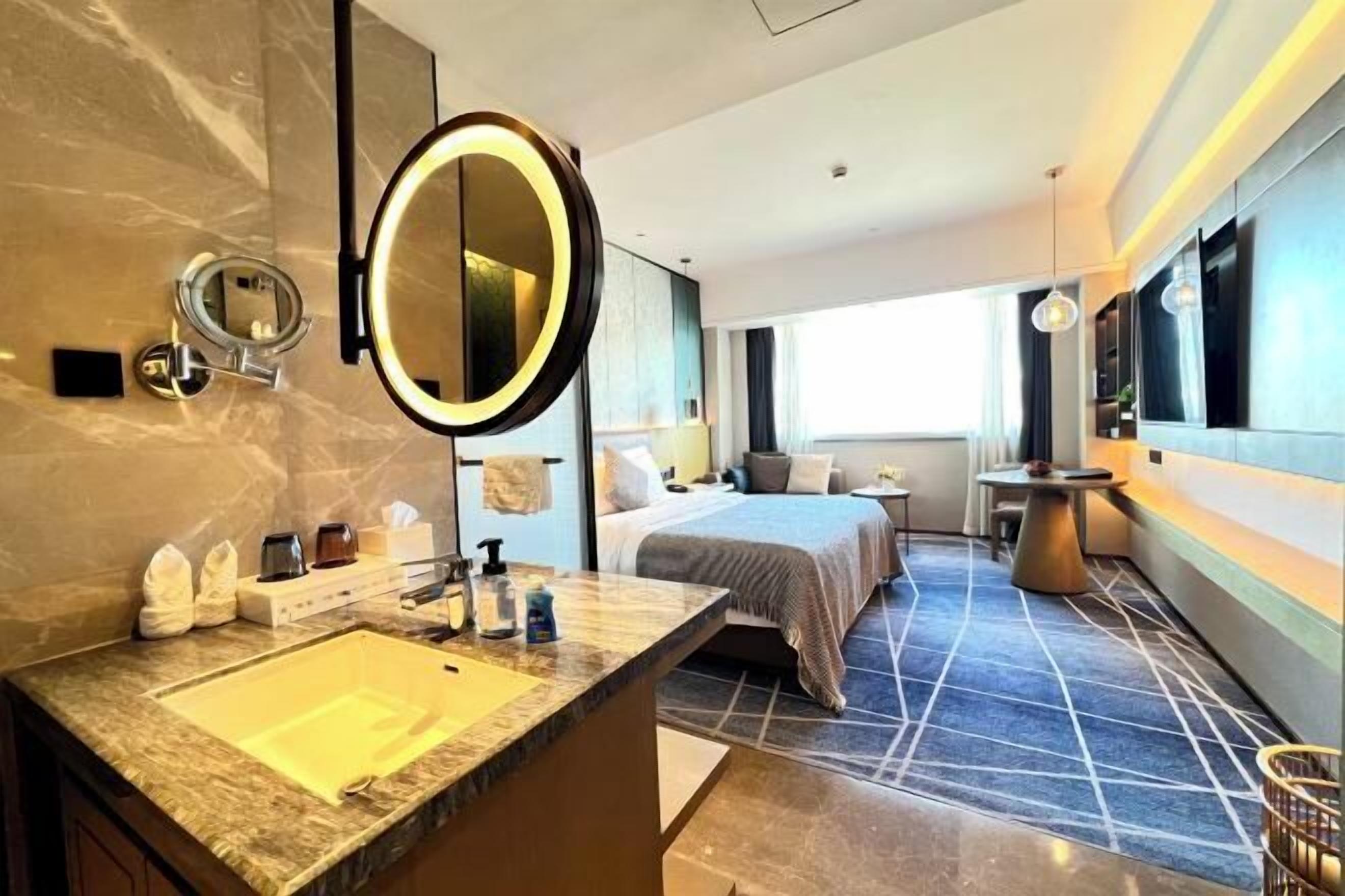 A modern hotel bathroom with a marble sink, a large mirror, a bed, and a small table.