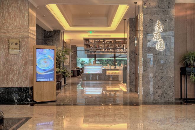 Een hotel lobby met een receptiebalie, een digitaal scherm en uitzicht op het restaurantgedeelte.