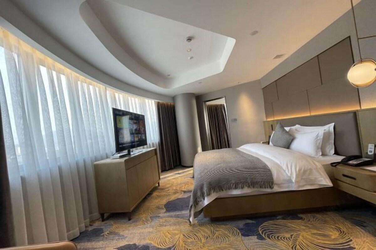 Une chambre d’hôtel moderne dotée d’un grand lit, d’une télévision à écran plat, d’une table de chevet et d’une grande fenêtre avec des rideaux.