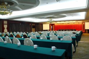 A conference room with rows of tables and chairs, a stage with a red backdrop, and a chandelier.