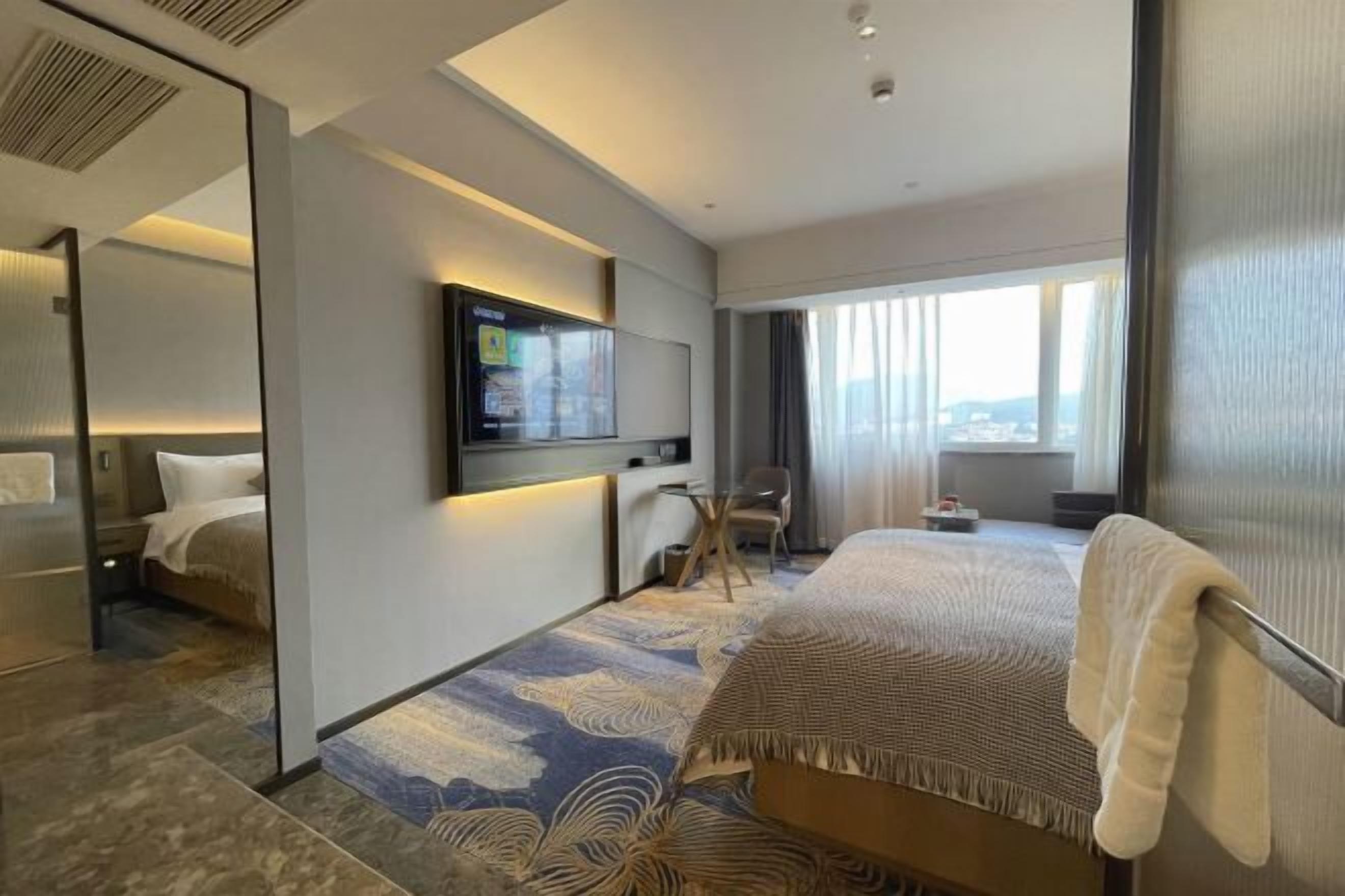 A modern hotel room with a large bed, a TV mounted on the wall, a desk, and a view of the city through the window.