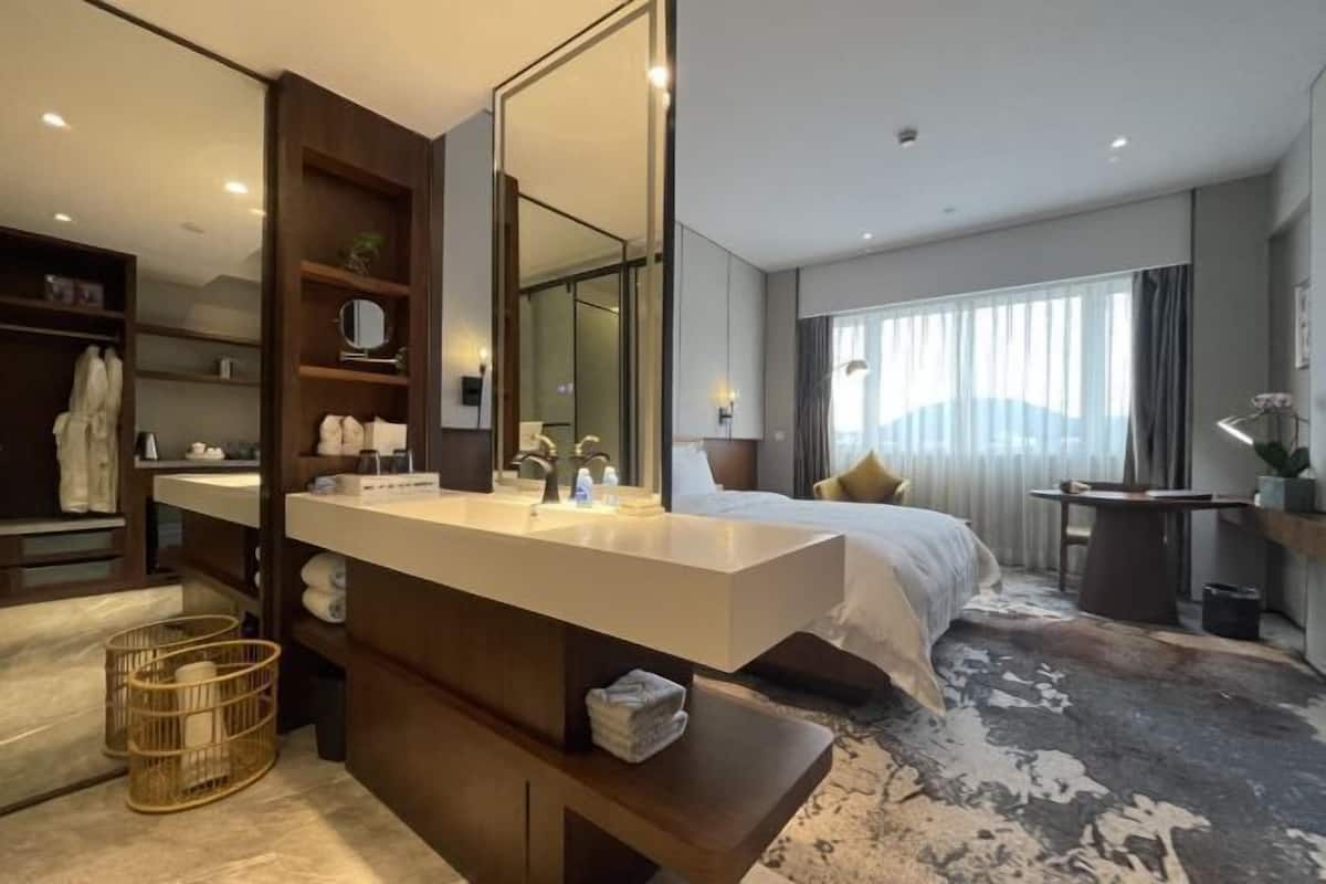 A modern hotel room with a large mirror, a wooden vanity, a bed with a white comforter, and a large window with curtains.