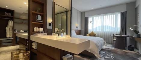 A modern hotel room with a large mirror, a wooden vanity, a bed with a white comforter, and a large window with curtains.