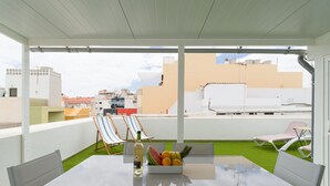 Eine Dachterrasse mit Tisch für zwei Personen, zwei gestreiften Liegestühlen, einer Obstschale und einer Weinflasche.