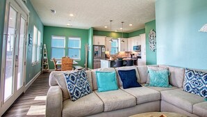 A living room with a beige sectional sofa, teal walls, and a kitchen in the background.