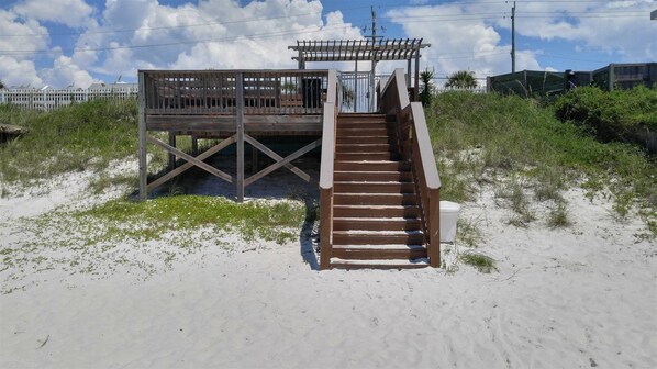 A wooden staircase leading to a beachside structure with a railing, surrounded by sand and sparse vegetation.
