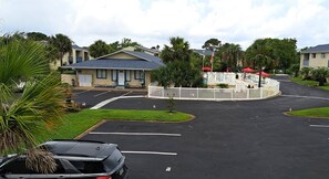 Un parking avec un bâtiment en arrière-plan, entouré de palmiers et d’une zone délimitée par une clôture, où se trouvent des parasols.