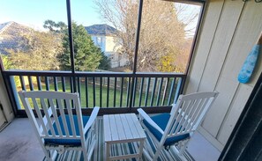 Un balcon avec deux chaises blanches et une petite table, donnant sur une cour arrière avec une clôture en bois et des arbres.