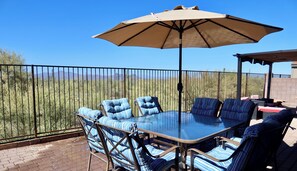 An outdoor patio with a glass table and striped chairs, a beige umbrella, and a view of a desert landscape. - Relax with Sonoran Nature Preserve views plus bike trails & several golf courses (Marana)