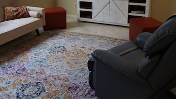 Una sala de estar con un sofá color beige, un sillón reclinable gris, un reposapiés rojo, un televisor sobre un mueble blanco y una alfombra estampada.