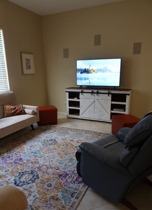 A living room with a beige sofa, a gray recliner, a red ottoman, a TV on a white entertainment unit, and a patterned rug. - Relax with Sonoran Nature Preserve views plus bike trails & several golf courses (Marana)