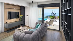 Ett modernt vardagsrum med en grå soffa, en platt-TV och utsikt över en pool och havet genom en skjutdörr i glas.