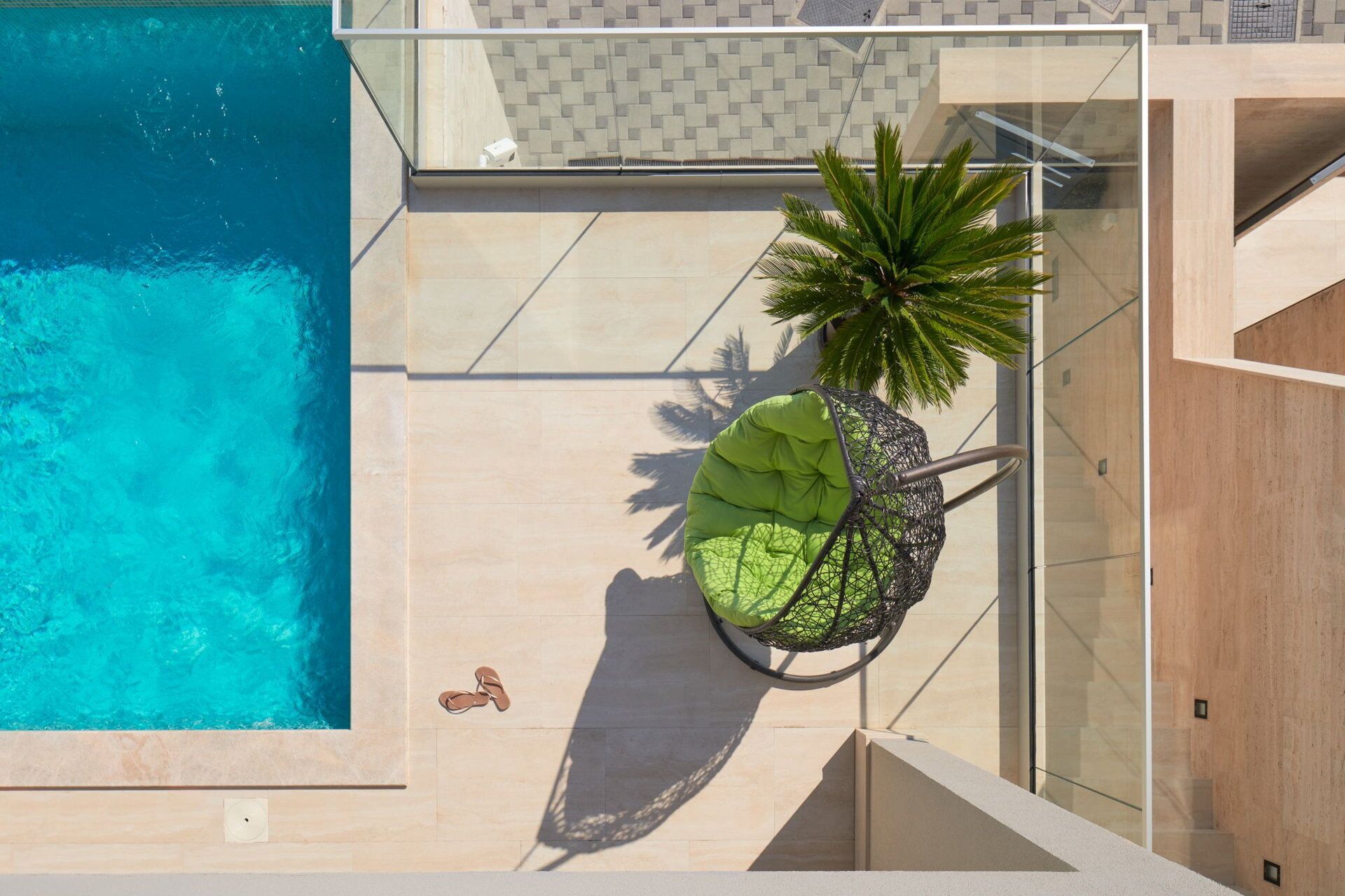 A rooftop pool with a glass balustrade, a green cushioned chair, and a potted palm plant.