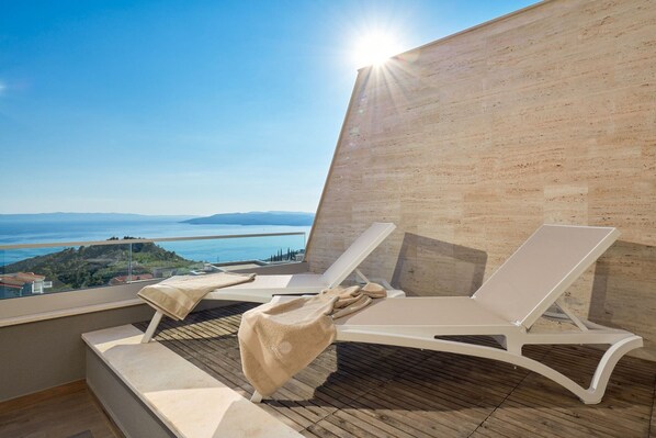 A balcony with two white lounge chairs and a small table, overlooking a scenic view of the sea and distant hills.