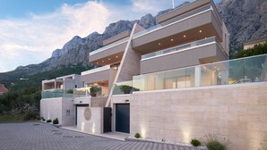 A modern multi-story building with a prominent glass facade, a geometric design, and a mountainous backdrop. - Belmare Villas Complex (Makarska)