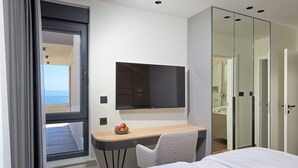 A modern bedroom with a bed, a desk with a chair, a flat-screen TV, and a view of the sea through a window. - Belmare Villas Complex (Makarska)