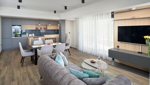A modern living room with a grey sofa, a coffee table, and a TV. There is a dining area with chairs and a kitchen in the background. - Belmare Villas Complex (Makarska)