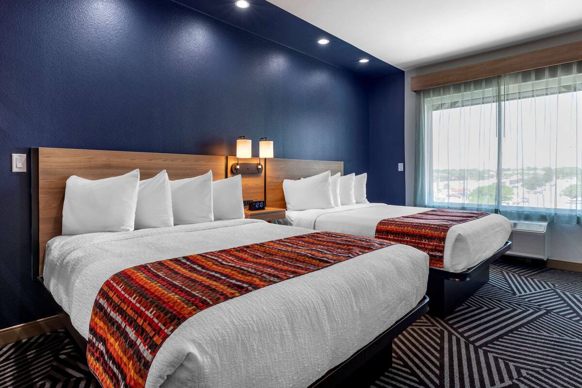 Een hotelkamer met twee bedden, een groot raam met gordijnen, en een hoofdeinde met wandlampen.